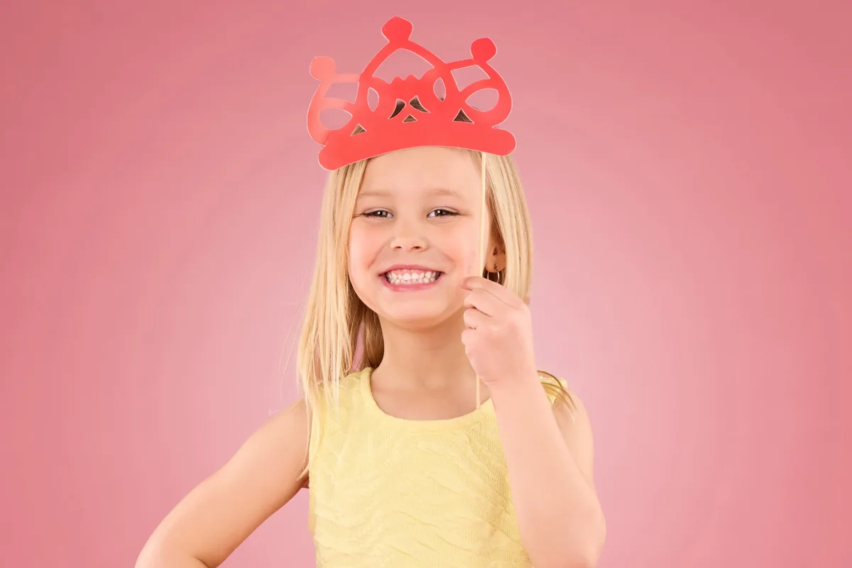 Smiling girl holding a red paper crown on a pink background